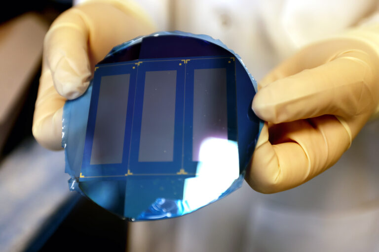 A pair of yellow gloved hands hold a blue wafer, which looks like a glossy, flat circle. Three gold rectangles are arranged vertically on the wafer. Each rectangle is framed by a thin, gold line.